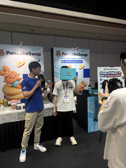 Two young men at a PancakeSwap booth, one holding up a blue branded sign while being photographed.