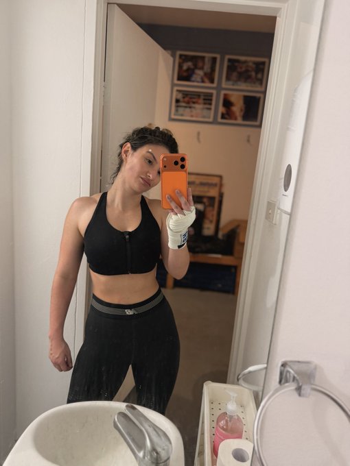 A mirror selfie of a woman wearing a black sports bra and black leggings, holding an orange iPhone in what appears to be a bathroom, with framed photos visible in the background.