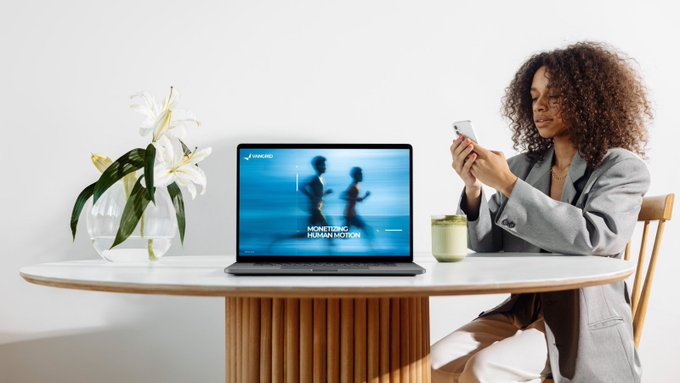 A woman with curly hair in a gray blazer smiling at her smartphone while sitting at a wooden table with white lilies, a matcha drink, and an open laptop displaying 'MONITORING HUMAN MOTION'.