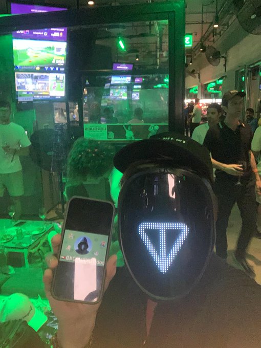A person in a sports bar holding up a phone displaying a friend request notification, standing next to another person wearing a black LED mask with a glowing triangle design.