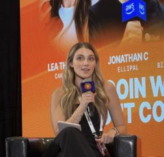 A woman with blonde hair speaks into a microphone at a panel, with text on the screen behind her reading "LEA TH...", "JONATHAN... ELLIPAL", and "COIN W...T CO...".