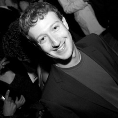 A black and white photograph of a smiling young man with curly hair and a blazer, taken in what appears to be a crowded setting.