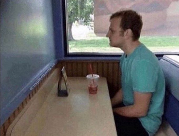 A side-profile view of a man in a teal shirt sitting at a diner table with a drink and a phone holder, looking out a window, with a '@cryptomemes.us' watermark.