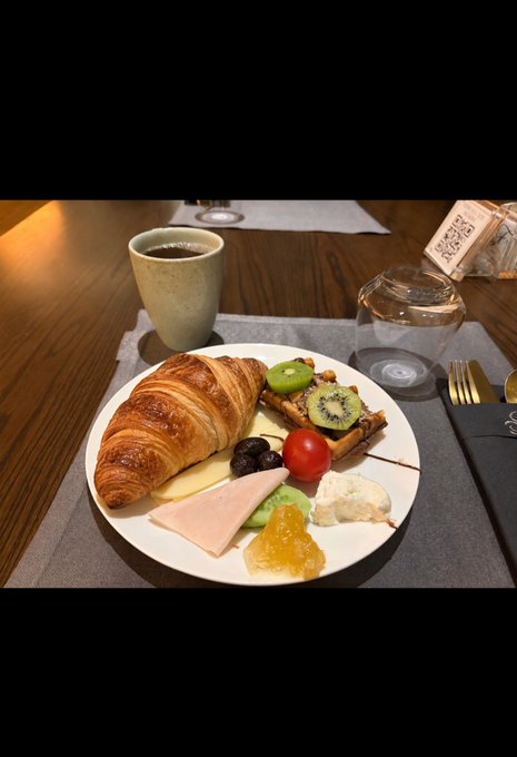 A buttery croissant takes center stage on a breakfast plate alongside fresh kiwi, a cherry tomato, dark berries, ham, cheese, and waffles with honey and jam, paired with a cup of coffee on a wooden table. The leisurely spread perfectly captures the relaxed, slow weekend vibe of the morning post.