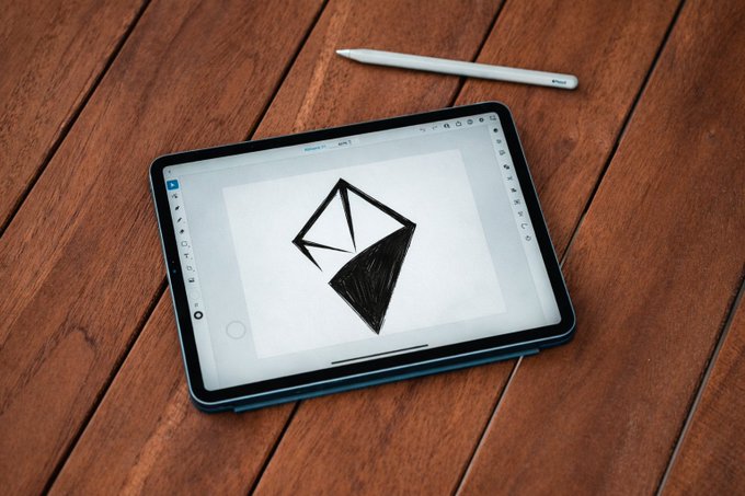 A tablet on a wooden surface displays a black digital sketch of an angular geometric shape alongside a white stylus, symbolizing the creative intersection of AI-generated content and blockchain-based digital asset ownership that AINFT enables.