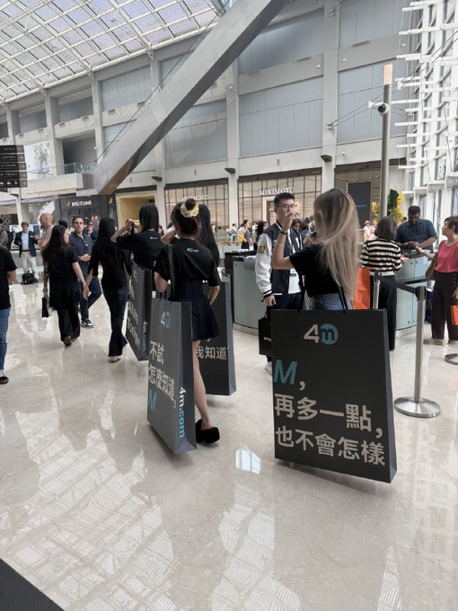 A line of young, stylish women wearing matching black outfits hold up 4M-branded signage in a modern building lobby, with visible text reading "4M," "我知道," and "再多一點,也不會怎樣," — a setup that playfully supports the post's joke about the crypto exchange looking more like a male model recruitment event.