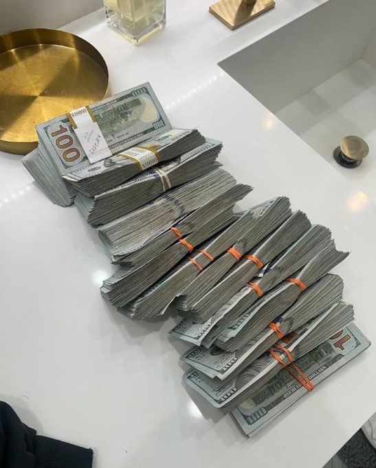Large stacks of $100 bills secured with orange and yellow bands piled on a white countertop near a sink and gold-colored plate.