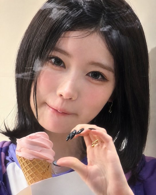 A young woman with dark, shoulder-length hair eats from a pink soft-serve ice cream cone, posing closely for the camera while wearing a ring on her finger.