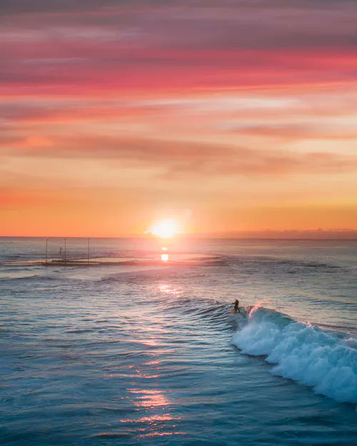 A scenic ocean view at sunset with vibrant pink and orange skies, showing a surfer riding a wave while an outrigger boat sits on the horizon.