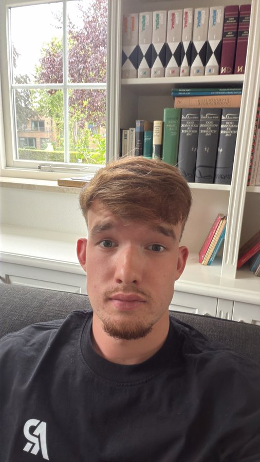 A young man with short brown hair wearing a black sweatshirt taking a selfie in front of a bookshelf with a window in the background.