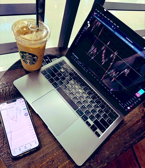 An iced Starbucks coffee sits beside a laptop and smartphone displaying active trading charts, embodying the "gm" morning routine of reviewing the markets before the day begins.