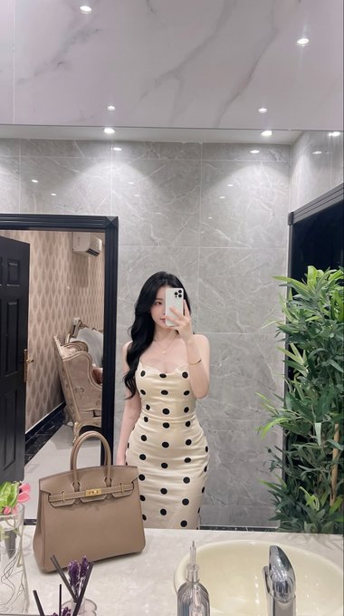 A young woman takes a mirror selfie in an elegant bathroom while wearing a cream-colored polka dot dress, with a tan Birkin-style handbag placed on the counter beside her.
