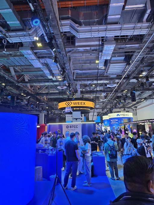 A busy indoor cryptocurrency conference featuring booths for WEEX, BTCC, MEXC, and OKX, with attendees networking under bright blue lighting and exposed industrial ceilings.
