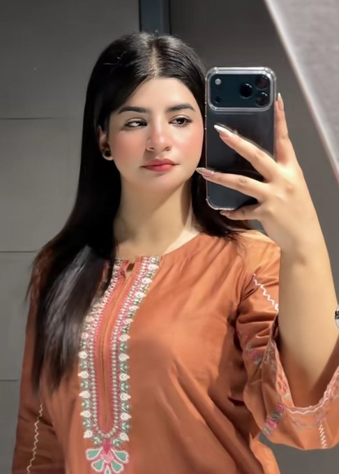 The image features a woman taking a selfie with her cell phone. The photo captures her face, as well as the reflection of the cell phone screen. She appears to be wearing an orange dress or outfit that stands out against the background. The caption provided suggests that she is sharing this moment on social media platforms like Twitter and Instagram.