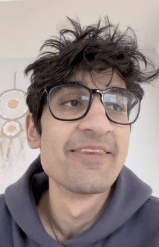 Close-up selfie of a young man with messy dark hair and black-framed glasses wearing a blue hoodie.