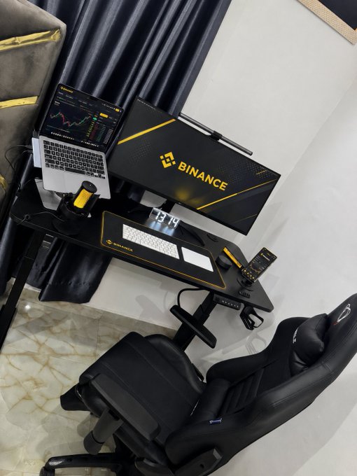 The image shows a Binance-branded desk featuring a cryptocurrency trading chart on a laptop, a digital clock reading 13:14, a keyboard, mouse, and gaming chair—reflecting a focused trader's workspace dedicated to building, learning, and staying connected in the crypto space.