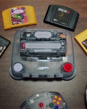 A transparent Nintendo 64 console is displayed alongside several game cartridges, including Zelda and Yoshi's Story, with a matching clear N64 controller visible at the bottom. The image showcases the see-through "Funtastic" edition of the console, with the post making a political statement about the owner of the device.