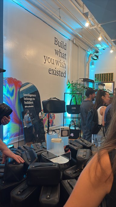A trade show booth displays the slogan 'Build what you wish existed' on the wall, with 'Intelligence belongs to builders' on a sign, surrounded by various tech bags and accessories.