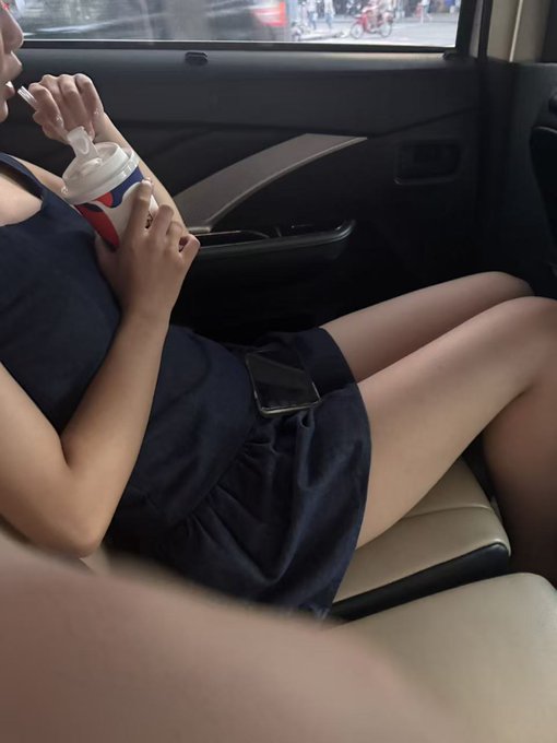 A person in a dark blue dress sitting in a car with their legs visible, drinking from a cup with a straw while holding a smartphone on their lap.