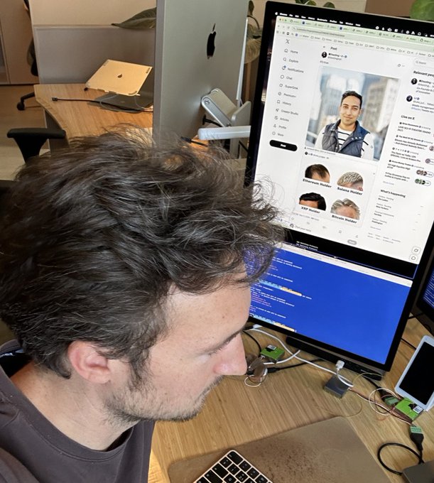 A side view of a curly-haired man working at a desk with two monitors; one displays the X (Twitter) social media platform.