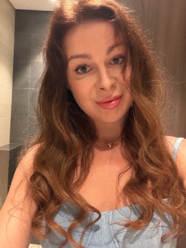 A selfie of a woman with long wavy brown hair wearing a light blue top and a small necklace, appearing to be in a bathroom setting.