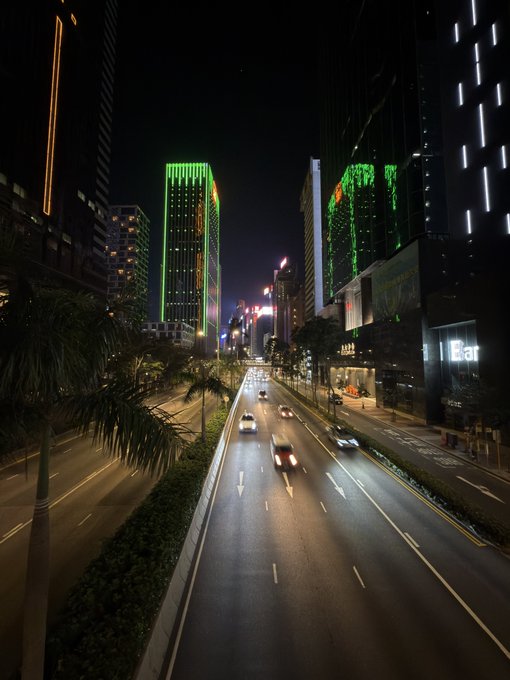 A nighttime aerial view of a city street with cars driving on a multi-lane road, flanked by palm trees and tall skyscrapers with green neon lighting on their facades.