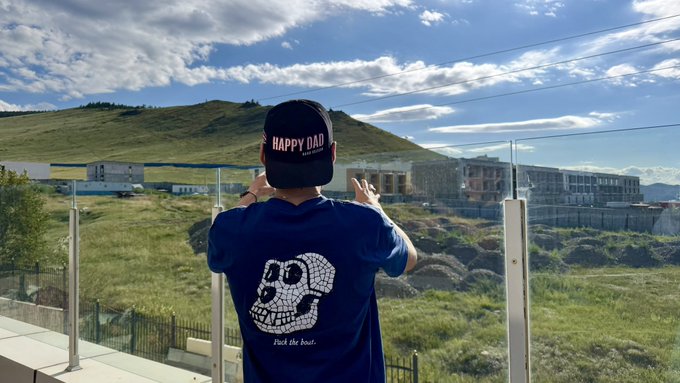 A person seen from behind wearing a black cap with 'HAPPY DAD' text and a navy blue t-shirt featuring a skull graphic and the phrase 'Pickle dick.' They appear to be taking a photo of a scenic landscape with hills, a lake, and apartment buildings.
