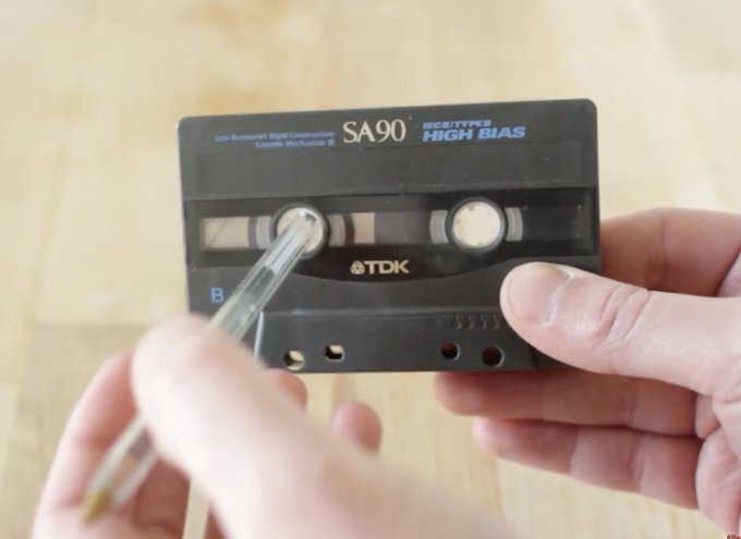 A close-up of hands holding a vintage TDK SA90 cassette tape labeled 'IECII/TYPE II HIGH BIAS' while a pen points to the magnetic tape inside.
