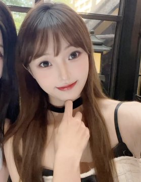 A young woman with long brown hair poses for a selfie, wearing a black choker and making a peace sign with her hand.