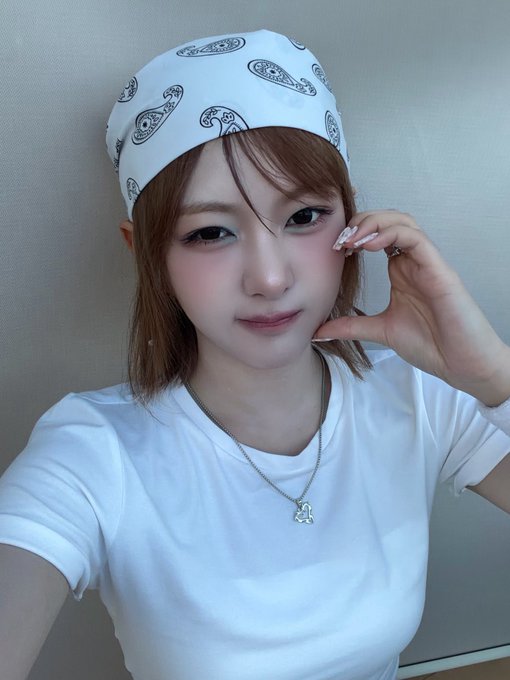A selfie of a young woman wearing a white paisley bandana, a white t-shirt, and a small teddy bear necklace, with long nails resting near her face.