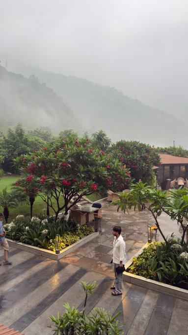A misty mountain resort scene with lush green hills, flowering red trees, and people walking on a wet stone pathway in the rain.