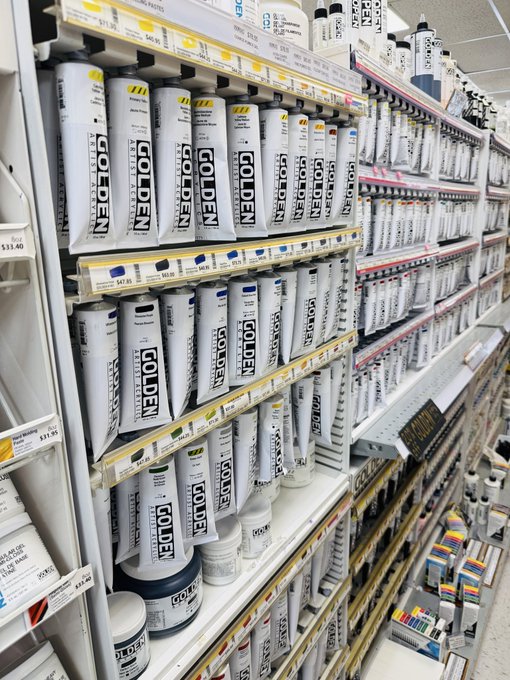 Store shelves stocked with rows of Golden brand acrylic paint tubes organized by color, with price tags visible.