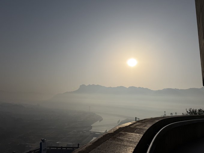 The image features a beautiful view of mountains, with a large sun setting behind them. The sky appears hazy due to the foggy conditions, creating an atmospheric scene. A person can be seen standing near the edge of a cliff or on a bridge in the foreground, adding depth and perspective to the image.