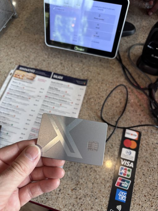 A hand holds a metallic silver credit card with a geometric X/K design and EMV chip in front of a Square point-of-sale terminal displaying a checkout screen. Visible on the counter are a menu showing 'SMASHED' and 'SALADS' sections, and a payment sticker decal featuring Discover, Mastercard, Diners Club, Visa, JCB, UnionPay, and American Express logos.