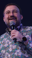 A bearded man in a patterned shirt holds a microphone and smiles while performing on a dimly lit stage.