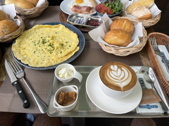 A delicious breakfast spread featuring a large omelet, baskets of fresh bread rolls, a cappuccino with intricate latte art, bacon, fried eggs, and small bowls of butter and jam on a café table.