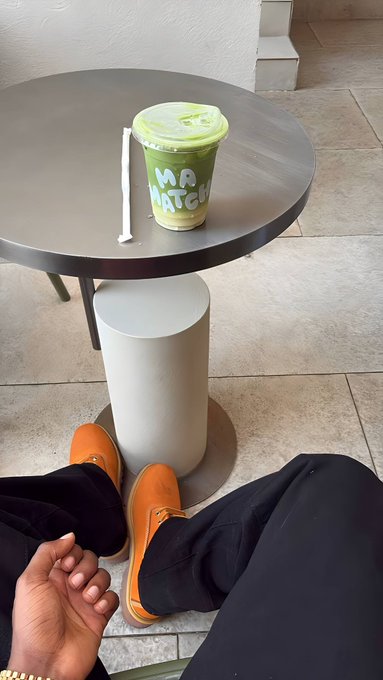 A first-person view shows someone wearing brown loafers and black pants, sitting at a round dark table holding a green matcha drink cup with 'MA MATCHA' printed on it.