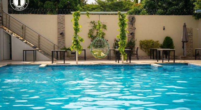 A resort or hotel swimming pool area featuring clear blue water, a hanging egg-shaped swing chair, tables, and lush green plants against a beige wall.