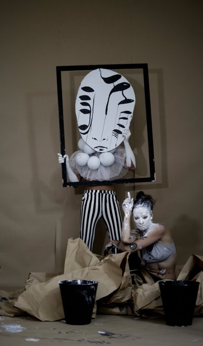 An avant-garde art performance featuring a person holding a frame containing an abstract black and white face mask above their striped legs, while another white-painted figure crouches beside them near black buckets.
