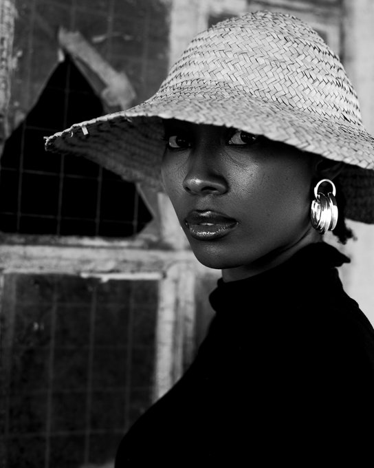 A striking black-and-white portrait captures a woman wearing a wide-brimmed straw hat and silver hoop earrings, her face partially shaded as she looks over her shoulder.
