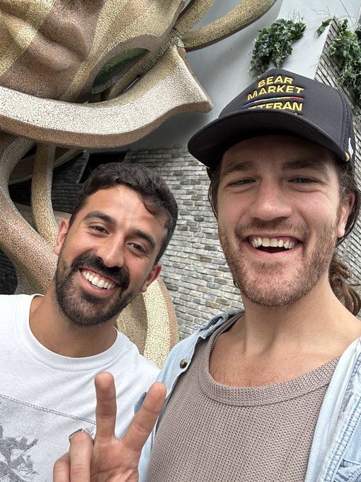 The image features two smiling men who are posing next to a large sculpture in front of a building, likely the Gear Market. They seem to be having a good time together, with one man making peace signs while the other looks back at him. The photo also includes a text that reads "Ran into my favorite XRP influencer in Miami. So glad he told me to buy before it goes to $10,000!" This is likely related to the gear market and Bitcoin exchange they are both talking about, as well as their interest in cryptocurrencies like XRP.