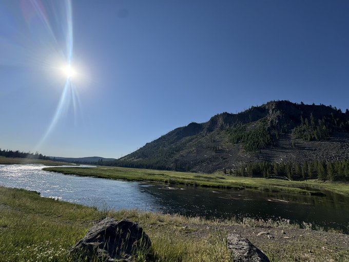 A scenic landscape photograph showing a river flowing through a grassy meadow with mountains and pine trees in the background, under a bright sun with lens flare in a clear blue sky.