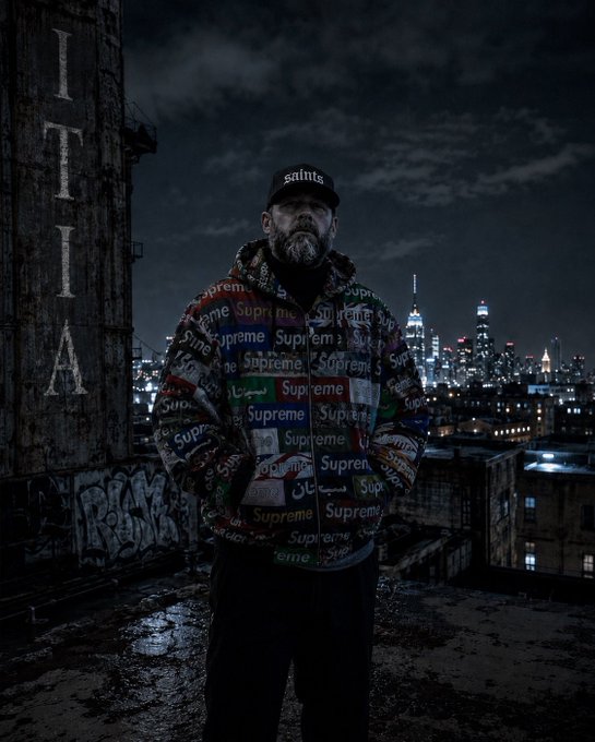 A man wearing a 'saints' cap and a colorful jacket patterned with Supreme logos stands on a rooftop overlooking a city skyline at night, near a brick wall reading 'ITALIA'.
