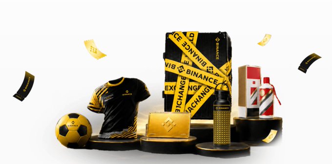 A Binance promotional product display featuring a soccer ball, black and yellow jersey, suitcase, yellow wallet, gold bar, and champagne bottle on black pedestals.