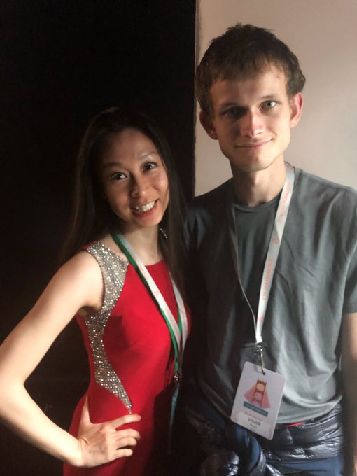 The image features a man and woman standing next to each other with smiles on their faces. The woman has long hair and is wearing a red dress, while the man has short brown hair and is wearing a gray shirt. They are both smiling at the camera, creating an endearing scene.