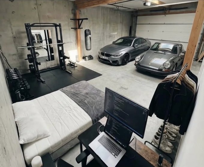 A combined living and garage space featuring a bed with a laptop and monitor setup, a weight rack with dumbbells and a punching bag, alongside two parked cars including a gray Porsche.