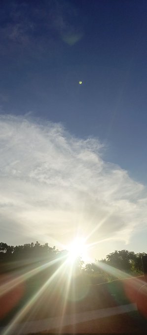 A bright sun shines low in the sky, creating lens flares through scattered clouds above a tree line, with a small celestial object visible high in the blue sky.