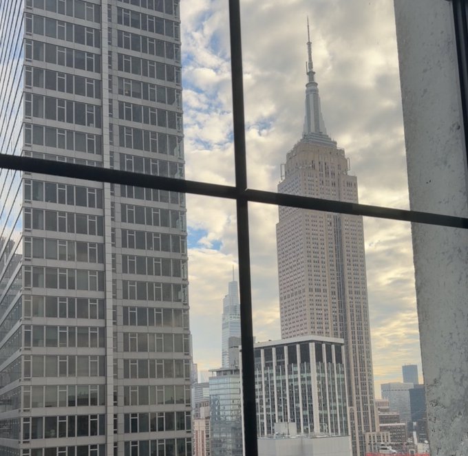 View of the Empire State Building through a multi-paned window with adjacent office skyscrapers visible under a cloudy sky.