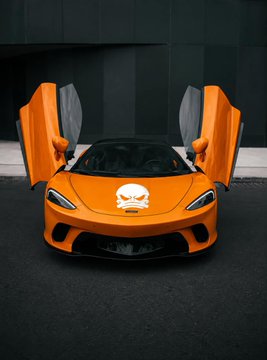 An orange sports car with its doors opened upward in a butterfly configuration, featuring a white skull logo on the hood, parked in front of a dark modern building.