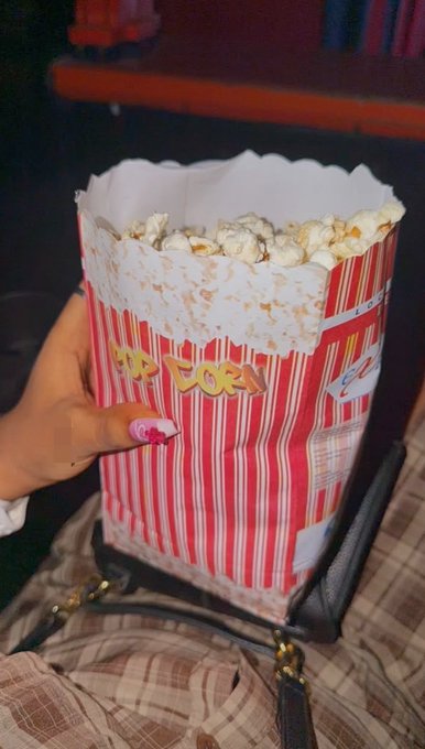 A hand with painted nails holds a red and white striped popcorn container filled with popcorn, with 'POP' partially visible, positioned in front of a black bag.